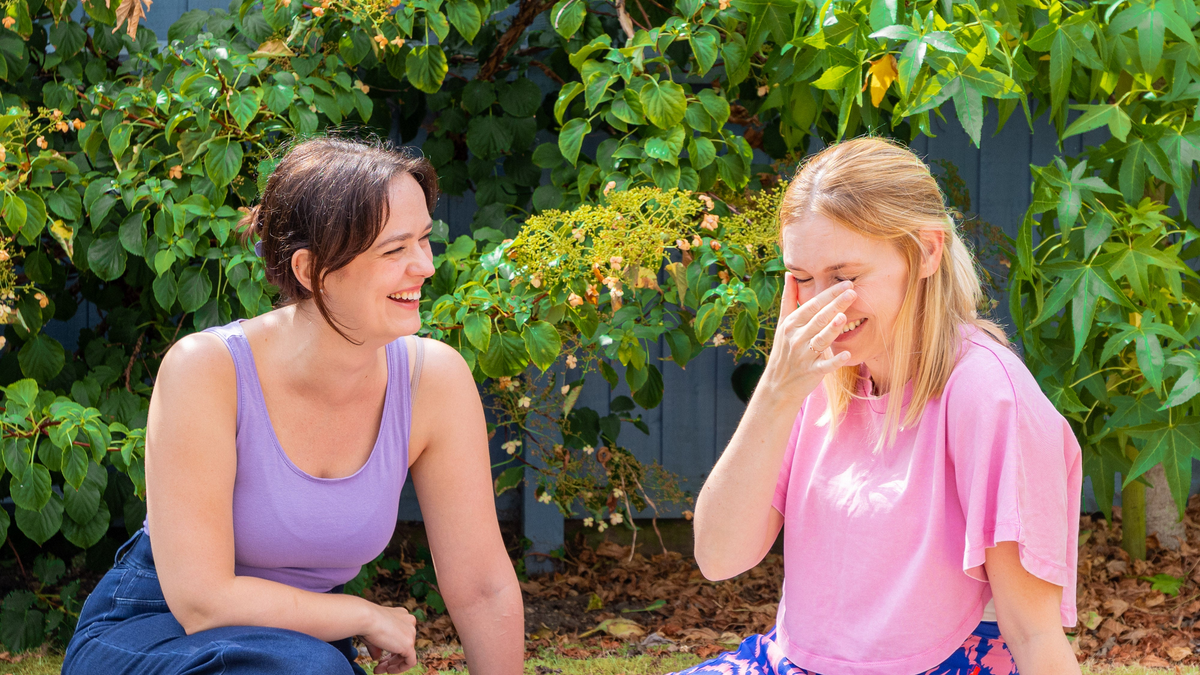 Two women sitting outdoors on the grass in front of a garden fence, smiling and laughing. One woman wears a lavender tank top and dark blue pants, while the other wears a pink t-shirt and a blue patterned skirt.