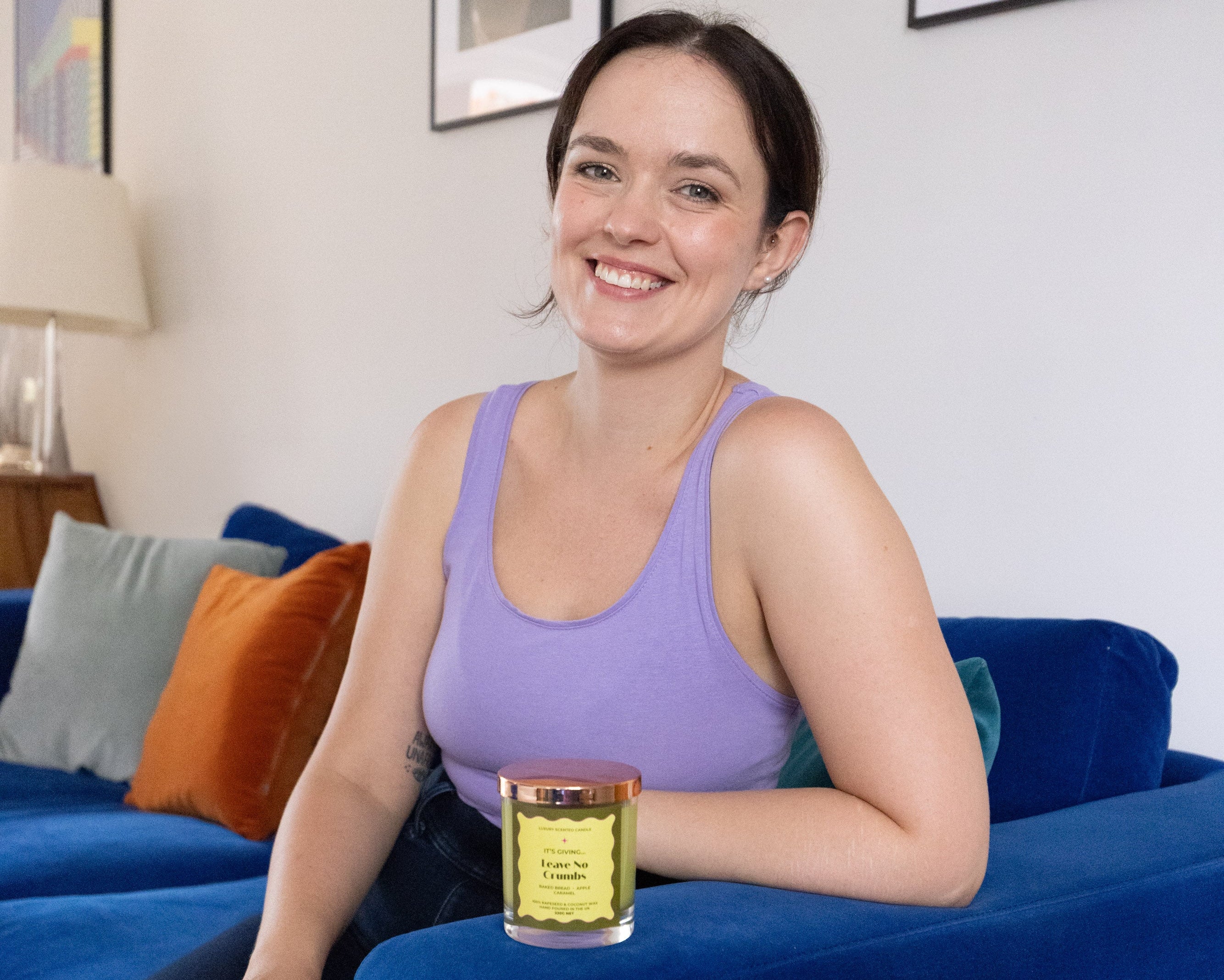 A woman with dark hair tied back sits on a blue velvet sofa in a living room, wearing a lavender tank top and holding a gold candle jar with a clear glass container.